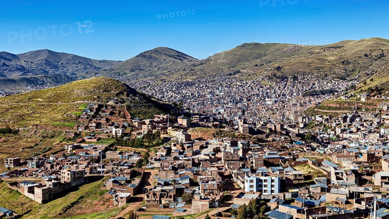 City view of buildings and mountains framed by green hills under a clear blue sky, view of the city of Puno in Peru