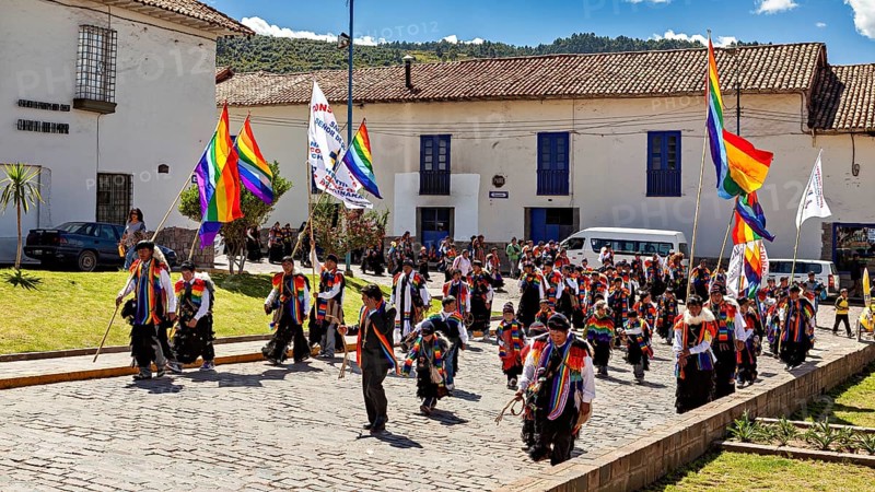 People with colorful flags march through a street with historic buildings under blue skies, traditional parade of festival groups in Cusco in Peru