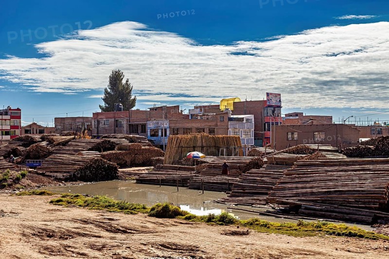 Rural cityscape with stacked wood and water surrounded by buildings under a cloudy sky, tropical timber warehouse near Puno in Peru