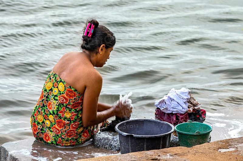 A woman is sitting by the river washing clothes with several buckets, a woman is washing at a lake near Sigiriya in Sri Lanka