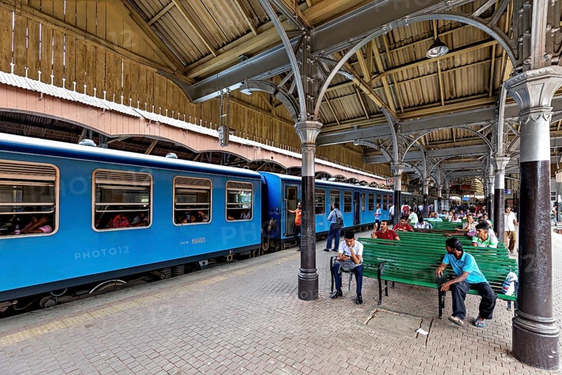 People sitting on green benches in the train station, blue train on the platform, covered structure, the station and platform with trains from Colombo in Sri Lanka