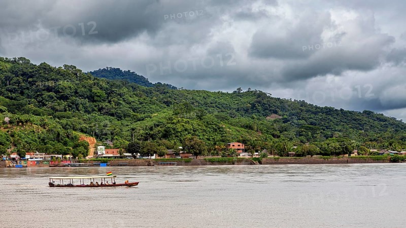 A boat moves on a river in front of a wooded hill and under a cloudy sky, people on boats near Rurrenabaque in the Amazon jungle in Bolivia