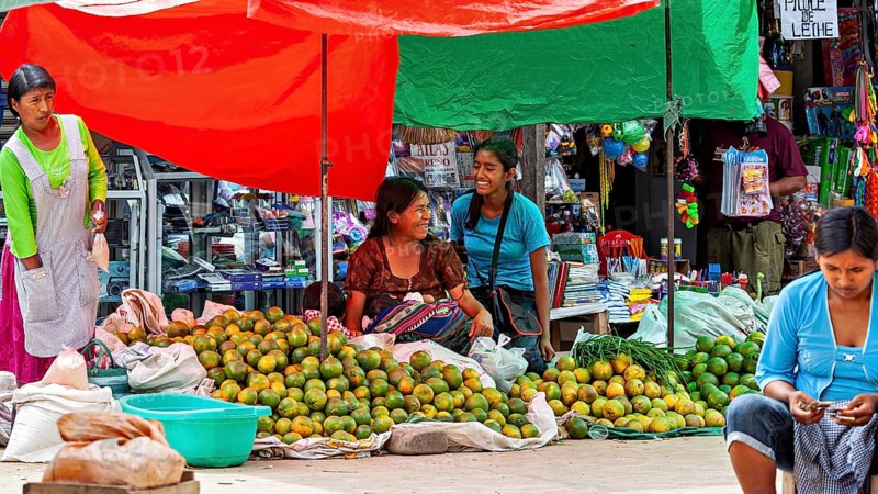 Two laughing woman sit in front of market stalls full of mangoes in a lively atmosphere, The Rurrenabaque market in Bolivia