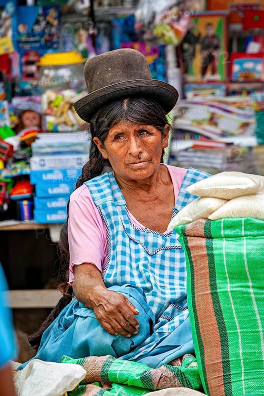 A thoughtful woman in traditional traditional costume sits in front of a colorful market stall, The Rurrenabaque Market in Bolivia