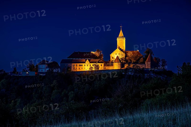 An illuminated castle is perched on a hill, surrounded by dark countryside and a deep blue night sky, The Leuchtenburg near Kahla in the Saale Holzlandkreis district in Thuringia