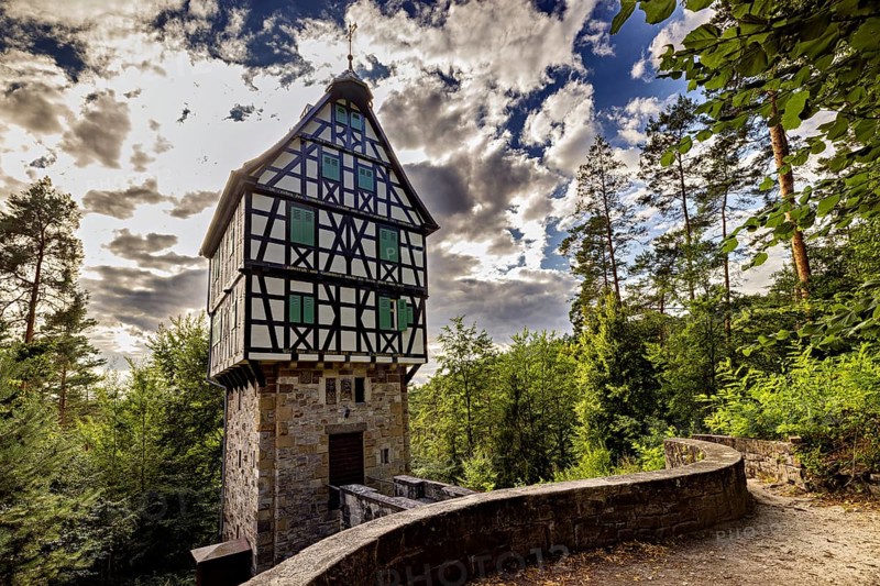 A tall, narrowly built half-timbered house on a stone base surrounded by dramatic skies with lush vegetation, The Herzogstuhl in the Saale Holzlandkreis district in Thuringia