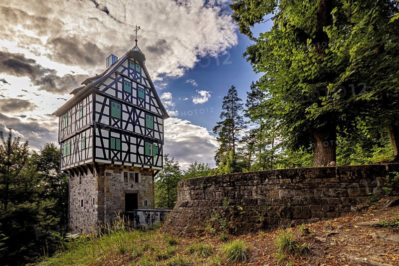 Half-timbered house on a stone base next to a stone wall in the forest, picturesquely illuminated by the sun with dramatic clouds, The Herzogstuhl im Saale Holzlandkreis in Thuringia