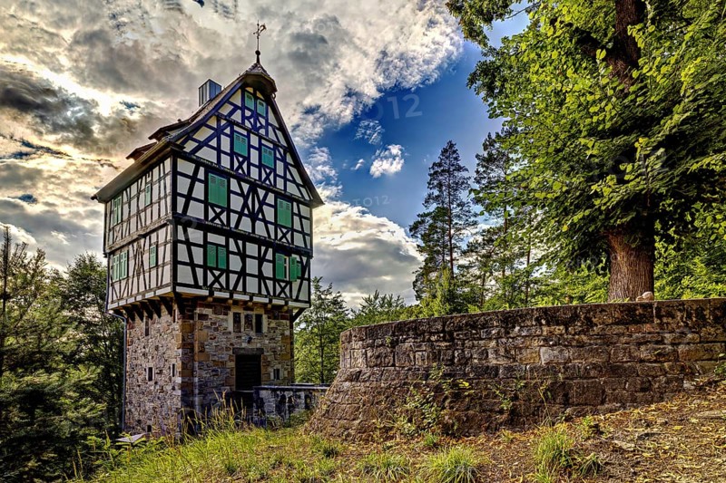 Half-timbered house on a stone base in the forest, surrounded by trees and a stone wall, under a dramatically cloudy sky, The Herzogstuhl im Saale Holzlandkreis in Thuringia