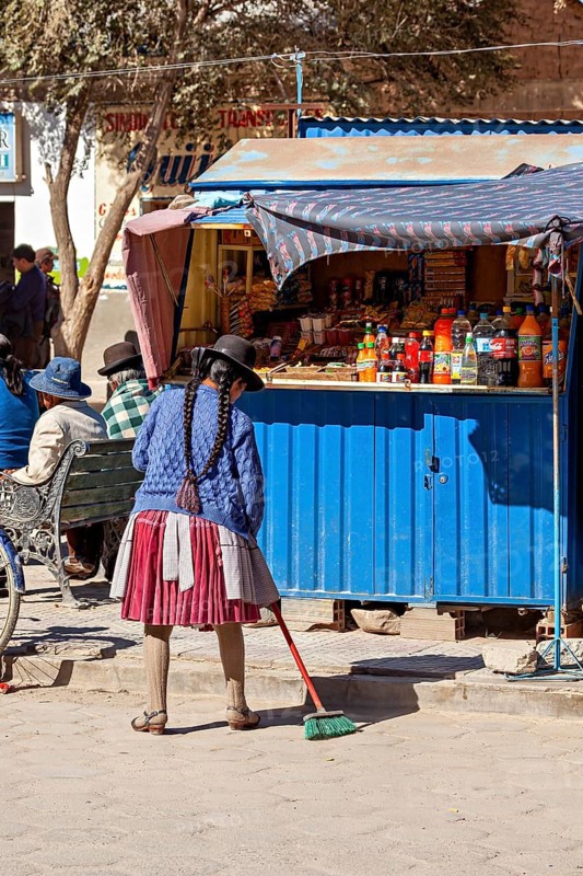 Woman in traditional dress sweeping in front of a blue stall on a busy street in the sunshine, Small shops in the town of Uyuni in the Salar de Uyuni in Bolivia