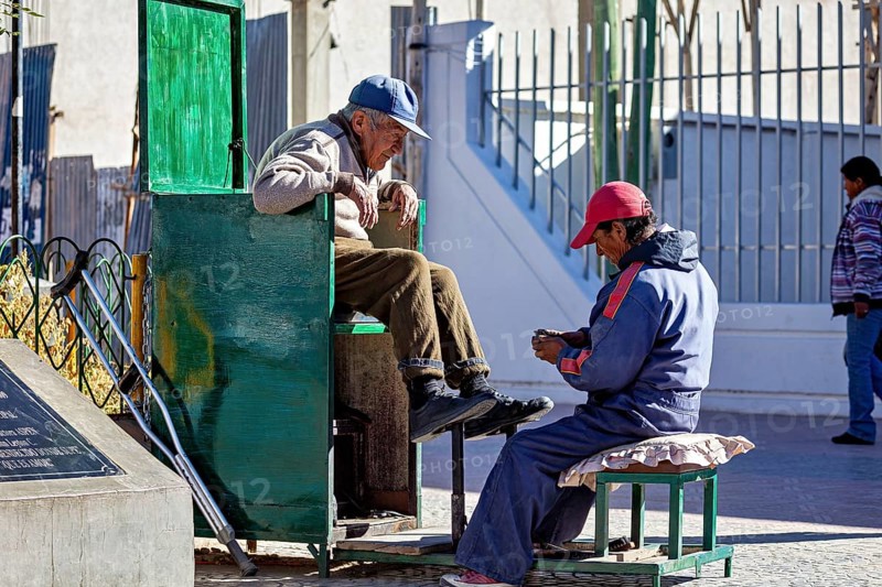 Shoe shiner concentrates on a man sitting on a green bench outdoors, show shiner in the city of Uyuni in the Salar de Uyuni in Bolivia