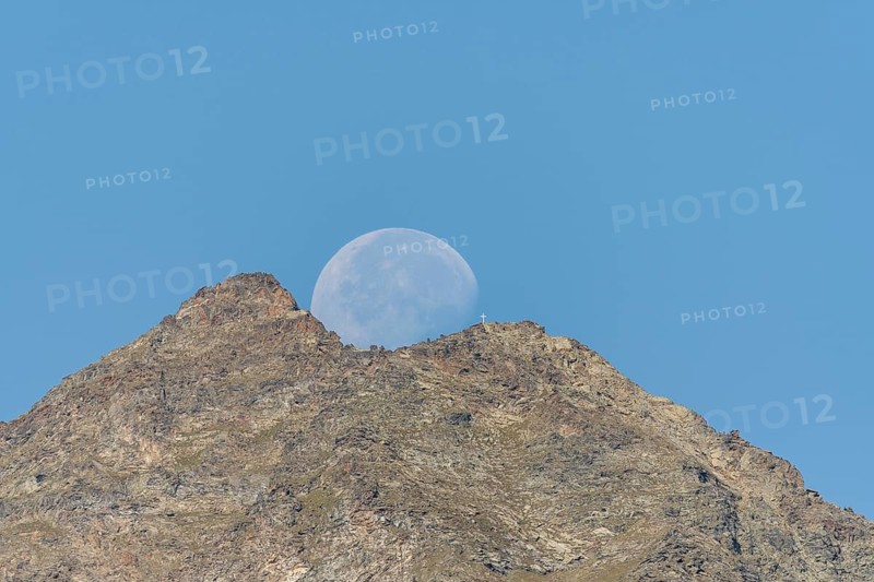 Majestic mountain peak stands tall against vibrant blue sky as large moon rises just behind it. The scene captures the quiet beauty of nature in the early evening light. Grachen, Viege, Valais, Switzerland
