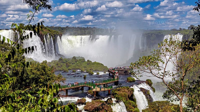 View of a lively waterfall and tourists on a bridge, surrounded by lush greenery and clouds, The Iguazu Falls between Argentina and Brazil