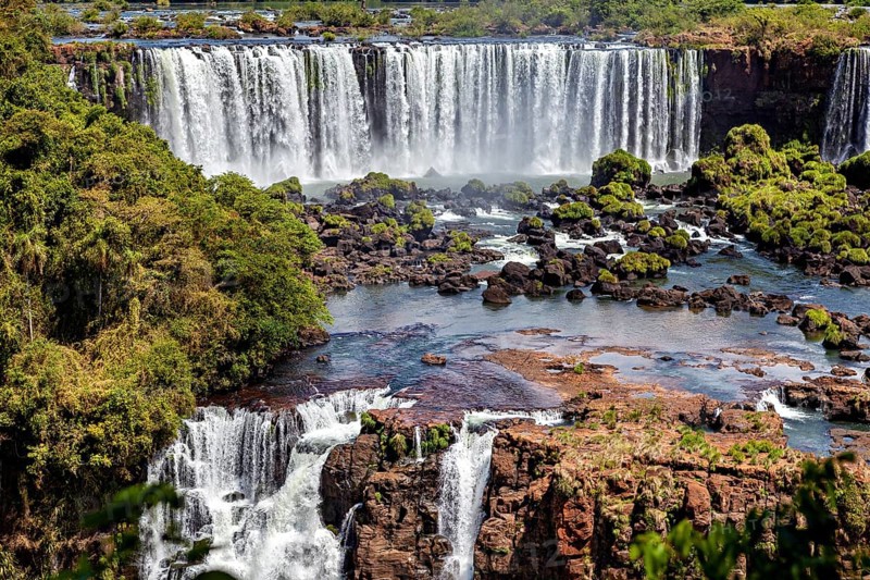 Waterfalls cascade over rocks into a wide river surrounded by dense forest, The Iguazu Falls between Argentina and Brazil
