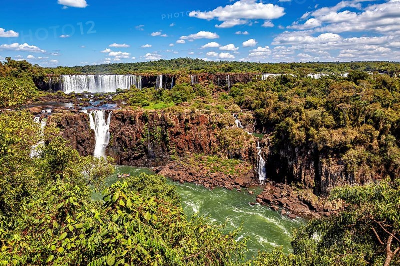 Waterfalls cascade into a river amidst lush vegetation and clear skies, The Iguazu Falls between Argentina and Brazil