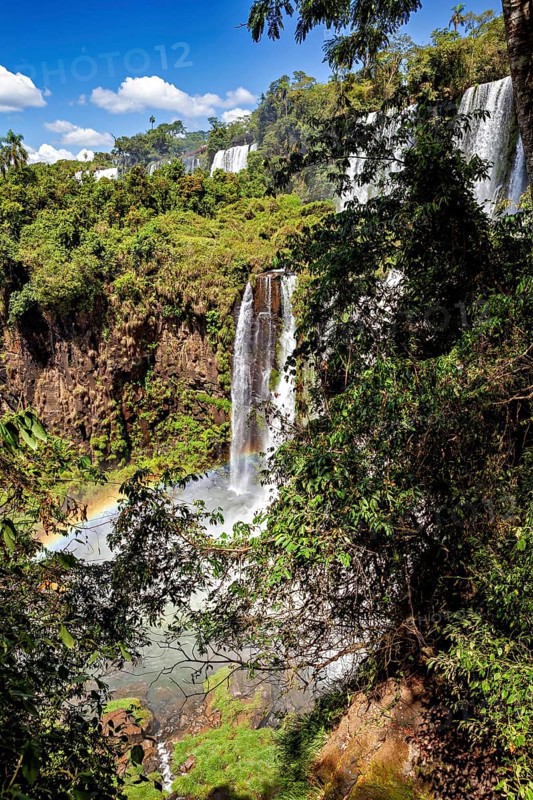 A narrow waterfall plunges through lush greenery and dense vegetation onto stony ground, The Iguazu Falls between Argentina and Brazil