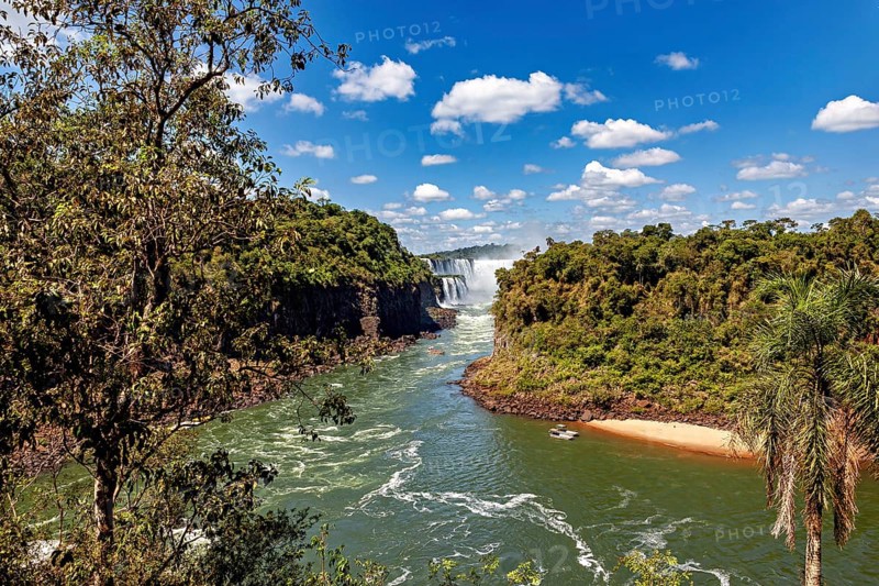 A wide river flows through a green landscape with jungle, while a distant waterfall adorns the background, The Iguazu Falls between Argentina and Brazil