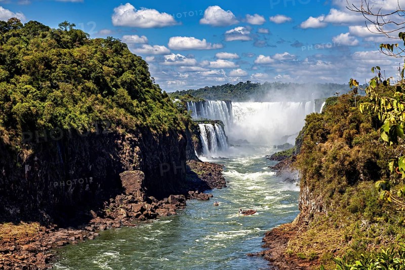 Majestic waterfalls flow between lush vegetation and steep rock faces under a clear sky with white clouds, The Iguazu Falls between Argentina and Brazil
