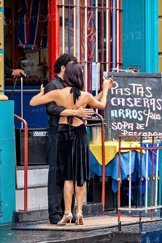 A couple dances tango in front of a colourful building, hugging each other, next to a menu board, The colourful city and tango district La Bocca in Buenos Aires Argentina