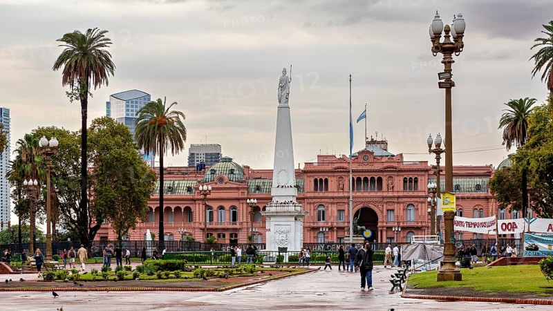 A monument on a square with a historic pink building and palm trees in the background under a cloudy sky, The presidential palace Casa Rosada in the city of Buenos Aires Argentina