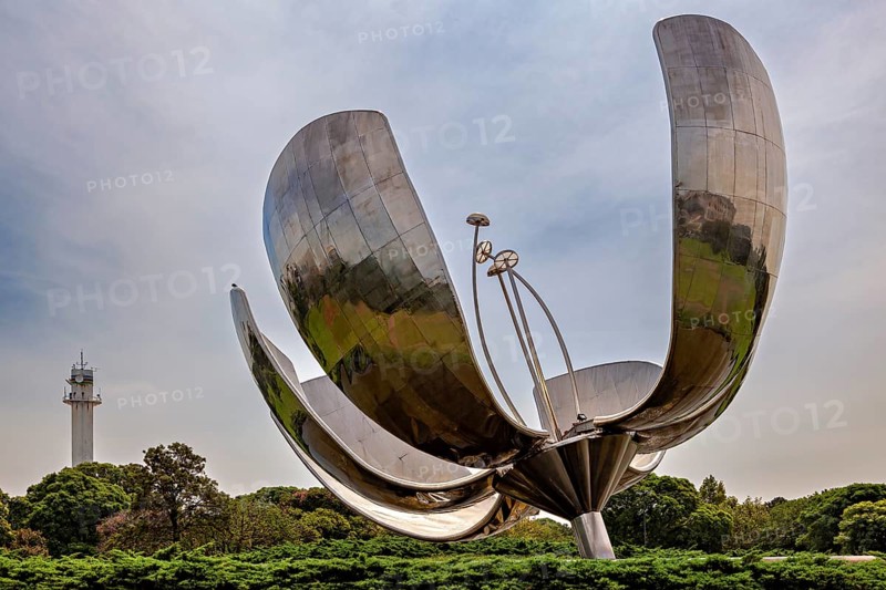 Metal flower sculpture next to a tower with a cloudy sky and green surroundings, The lotus blossom in Buenos Aires Argentina