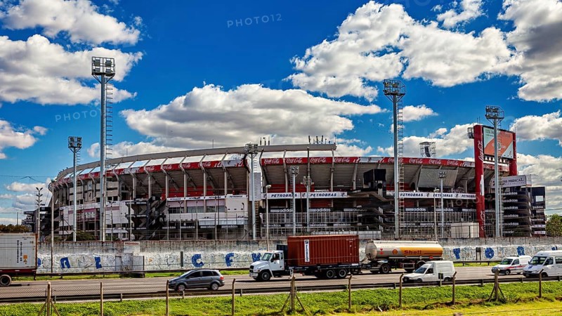 A large stadium with several stands and floodlights under a blue sky and passing vehicles, the River Plate football stadium in Buenos Aires Argentina