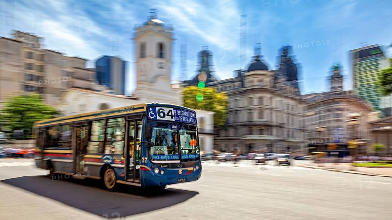 A colourful bus drives through a busy city with historic buildings and a blue sky in the background, bus route 64 in the city centre of Buenos Aires in Argentina