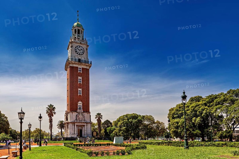 A clock tower surrounded by manicured gardens, palm trees and lanterns in a sunny park, The city of Buenos Aires in Argentina
