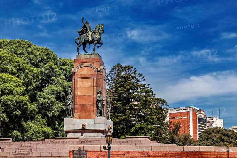 An equestrian monument on a pedestal surrounded by trees and buildings under a blue sky, The city of Buenos Aires in Argentina