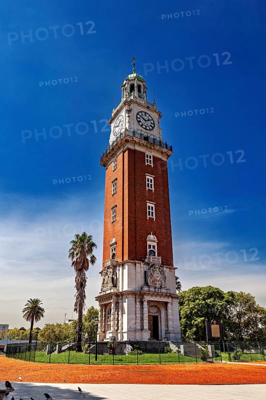A tall brick clock tower next to palm trees under a clear blue sky, The city of Buenos Aires in Argentina