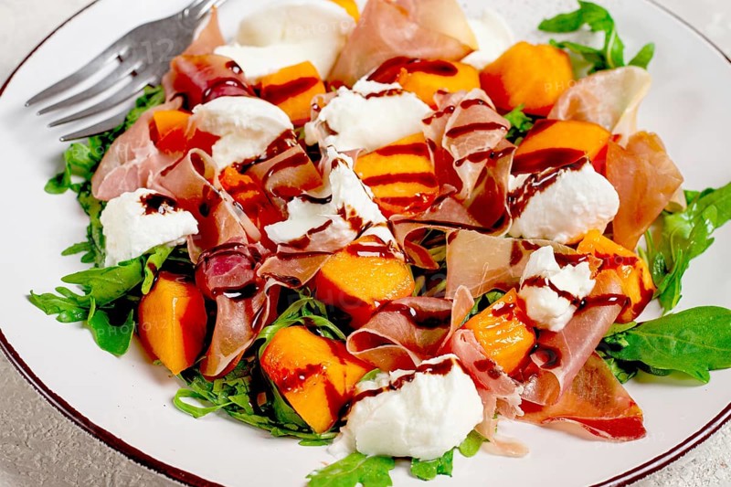 Salad with slices of ripe melon, prosciutto, mozzarella and fresh arugula, decorated with balsamic glaze