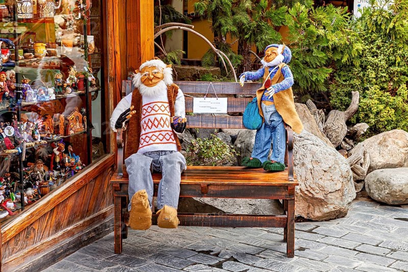 Two dolls sit on a bench in front of a shop window, surrounded by plants and wooden surfaces, dwarves and gnomes from Patagonia in South America