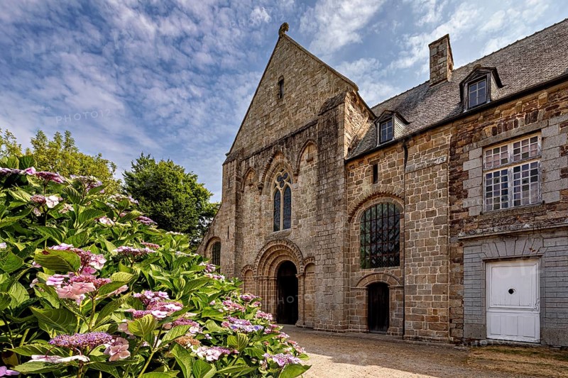 Historic stone church with colourful flowers in the foreground under a blue sky, The Abbey de La Lucerne in Normandy, France