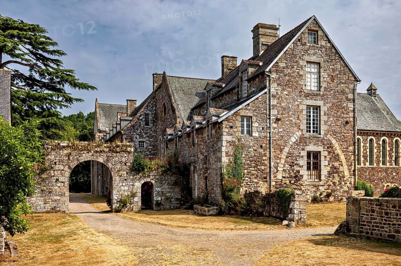 A historic stone building with archway and vegetation under a cloudy sky, The Abbey de La Lucerne in Normandy, France