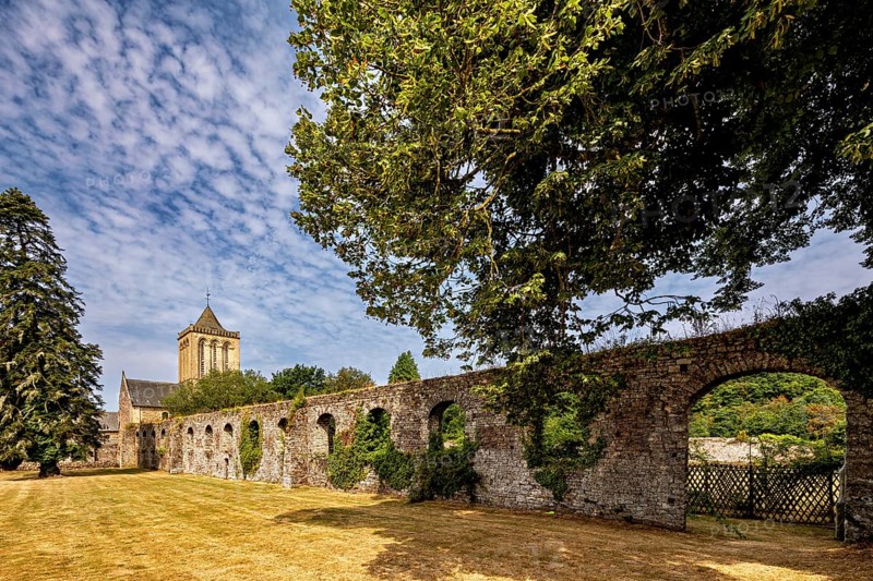 A vast stone wall next to a church, surrounded by trees under a clear sky, The Abbey of La Lucerne in Normandy, France
