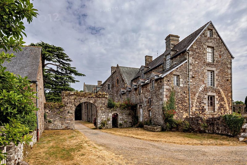 Historic stone building with an archway under a cloudy sky in a rural setting, The Abbey de La Lucerne in Normandy, France