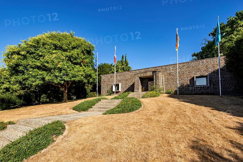 A modern building with flags, surrounded by trees and a dry meadow under a clear sky, German soldiers' cemetery from the Second World War near Huisnes-sur-Mer in Normandy in France