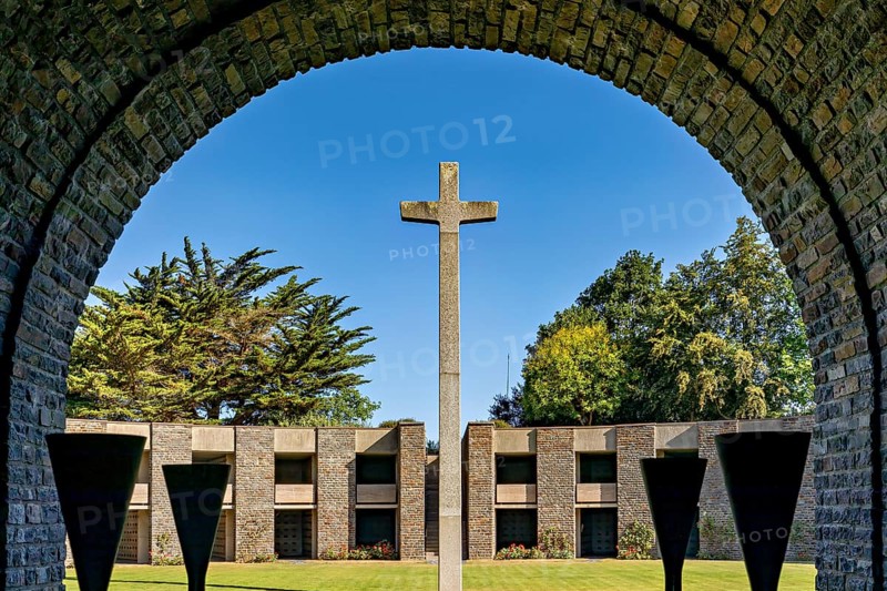 A stone cross stands in the centre of an arched passageway with a view of modern concrete architecture under a clear sky, German soldiers' cemetery from the Second World War near Huisnes-sur-Mer in Normandy, France