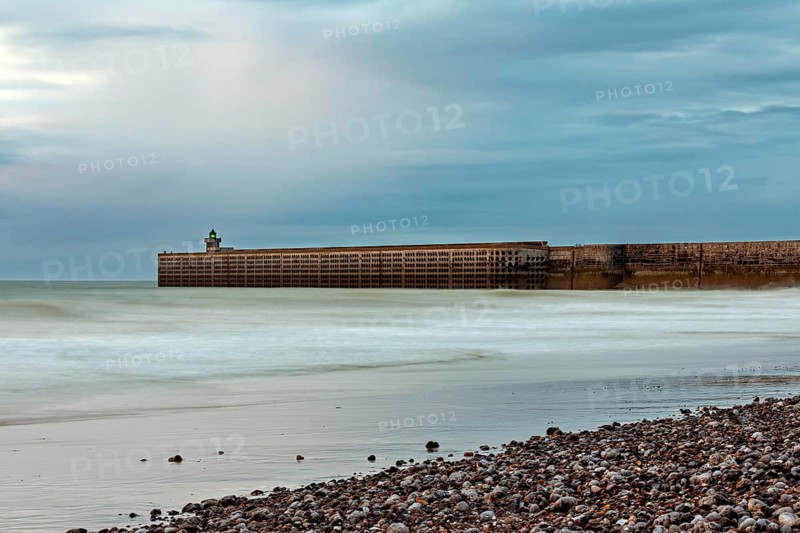 A breakwater juts into the sea, surrounded by a pebble beach and a cloudy sky, The pier and beach of Dieppe in Normandy, France