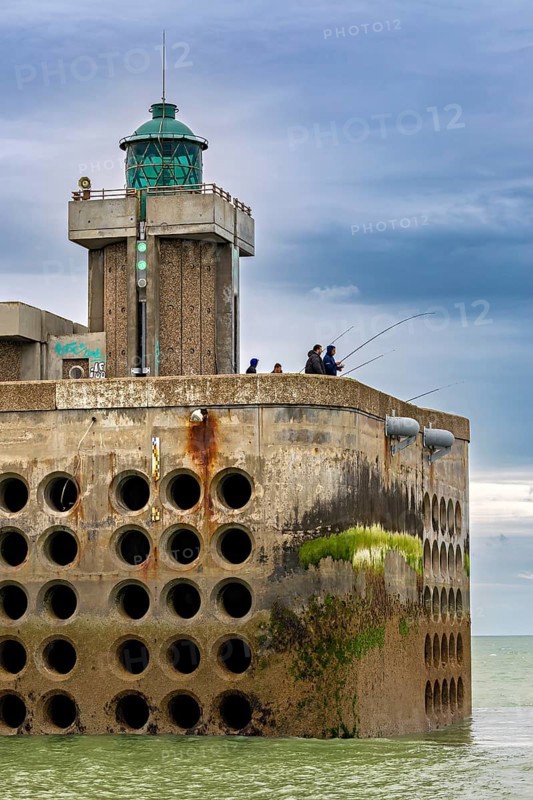 Concrete building with lighthouse and anglers at the water's edge, under a cloudy sky, The harbour pier of Dieppe in Normandy, France