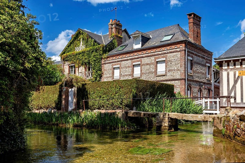 A brick house covered with climbing plants next to a clear body of water and lush vegetation, The historic mill village of Veules Les Roses in Normandy, France