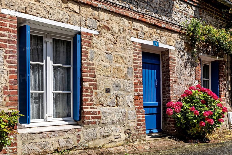 Stone house with blue shutters and door, flanked by hydrangeas under a bright summer sun, The historic mill village of Veules Les Roses in Normandy, France