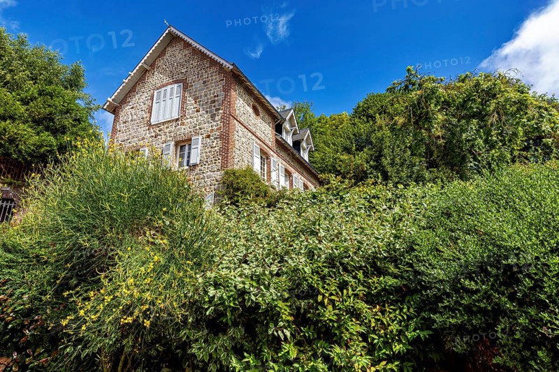 Single brick house behind green bushes and trees under a clear blue sky, The historic mill village of Veules Les Roses in Normandy, France