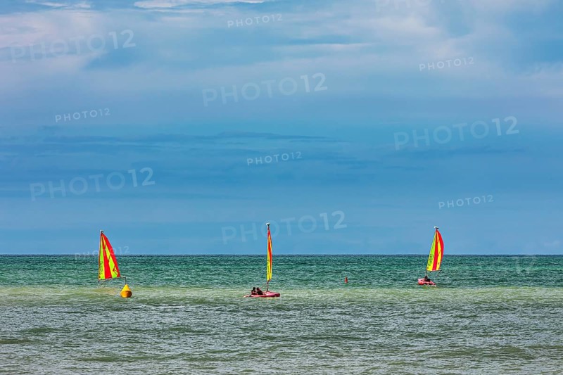 Three boats with yellow sails on the sea under a cloudy sky, surfers on the sea in Normandy, France