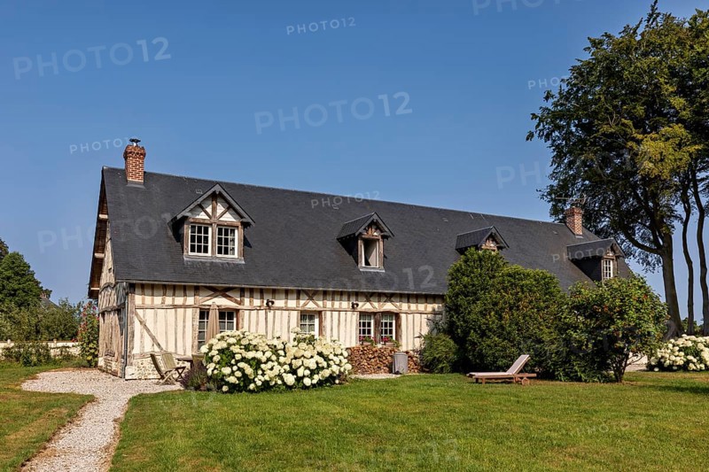 Half-timbered house with a dark roof surrounded by a well-tended garden and flowering shrubs under a blue sky, Historic houses in Normandy in France