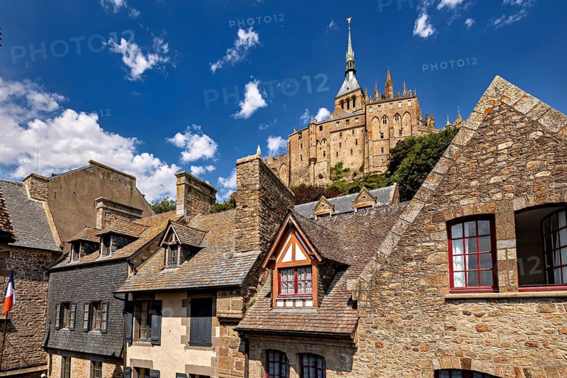 Stone and tiled roofs of ancient buildings with a dominant tower in the background under a clear sky, Mont Saint Michel in Normandy, France