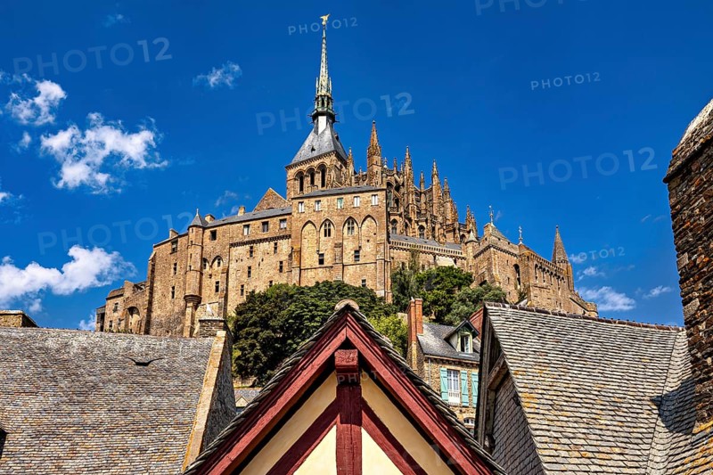 Medieval castle with a towering spire and rooftops against a clear blue sky, Mont Saint Michel in Normandy, France