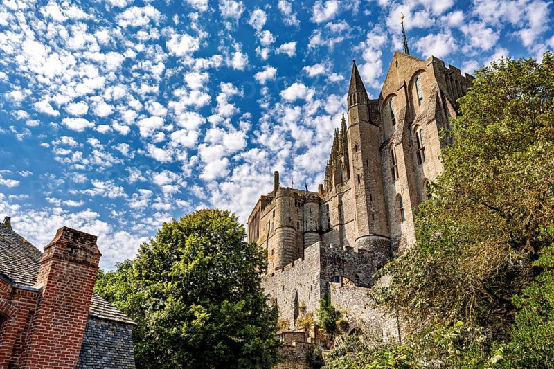Medieval castle rises dramatically into the sky, framed by green trees and old buildings, Mont Saint Michel in Normandy, France