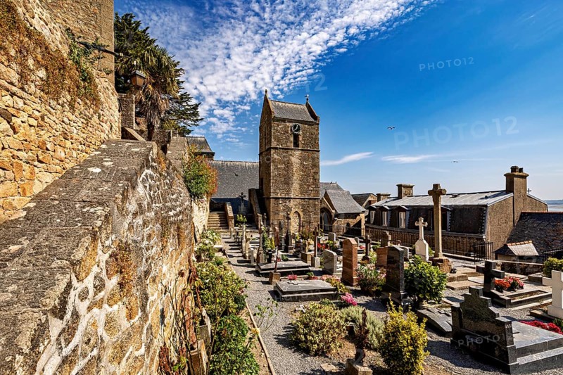 Historic cemetery with church and gravestones along a stone wall under a blue sky with clouds, The cemetery of Mont Saint Michel in Normandy, France