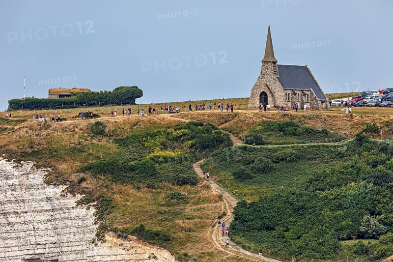 A small church stands on grassy cliffs with a path and walkers, under a blue sky, The church of Etretat in Normandy, France