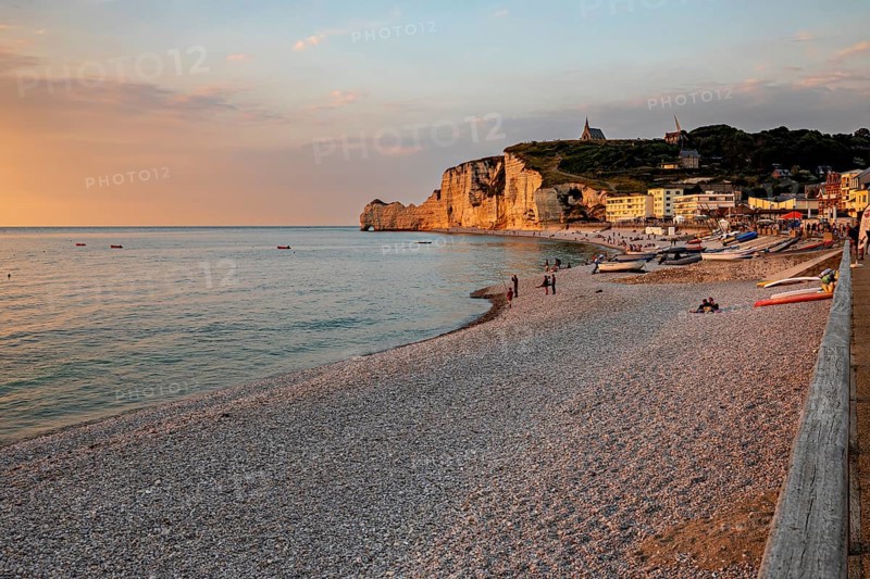 Beach with pebbles and boats, surrounded by rocks and a village, in the warm light of the sunset, The cliffs and alabaster coast of Etretat in Normandy France